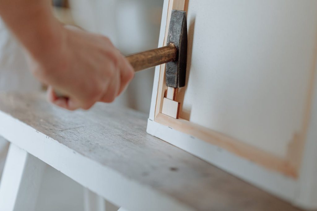 Detailed image of a hand using a hammer to work on a canvas frame in a workshop setting.